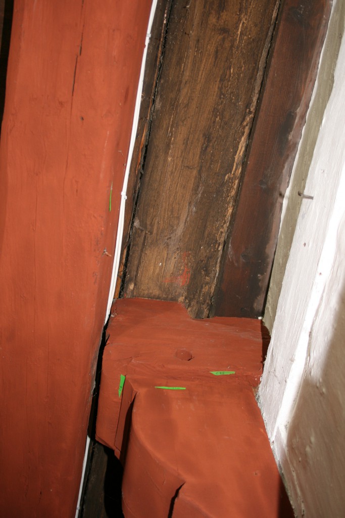 Den rødmalede bjælkeende, der stikker ud af væggen, er udskåret i en bølgelingende profil. Det kan man dog ikke se for den tværgårende røde bjælke.